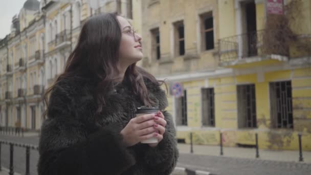 Стильная брюнетка кавказской внешности, девушка в солнцезащитных очках оглядывается по улице города и улыбается. Красивая молодая женщина пьет кофе во время поездки по Европе. Туризм, активный образ жизни, путешествия за границу . — стоковое видео