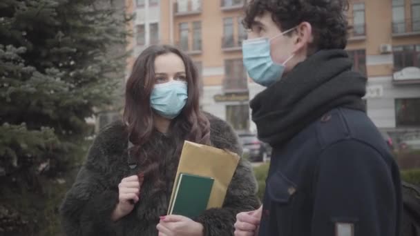 Retrato de una joven linda chica caucásica con mascarilla discutiendo noticias de coronavirus con su compañero de grupo. Estudiantes conscientes masculinos y femeninos con máscaras protectoras al aire libre y hablando. Cuarentena, pandemia . — Vídeos de Stock