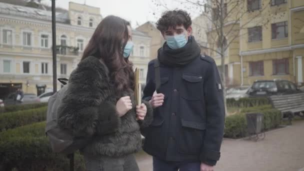 Porträtt av unga kaukasiska manliga college student i ansiktsmask prata med kvinnliga gruppkamrat utomhus. Pojke och flicka i skyddsmasker under karantänen. Covid-19, livsstil, hälsovård. — Stockvideo