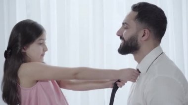 Orta Doğulu şirin bir kızın, babasının kravatını düzelttiği yakın plan. Mutlu küçük kız, gülümseyen iş adamının işe hazırlanmasına yardım ediyor. Aile, birlik, yaşam tarzı, bakım.