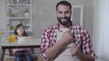 Önlüklü mutlu Orta Doğulu bir adamın portresi arka planda masada oturan bulanık küçük bir kız gibi poz veriyor. Evde kızına yemek yapan yakışıklı bir baba. Yaşam tarzı, aile, neşe.