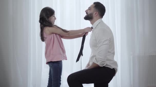 Oldalnézetben a közel-keleti kislányról, aki nyakkendőt köt a boldog apához. Aranyos lány fekete hajjal, segít a jóképű szülőknek felkészülni a munkára. Család, életmód, gondoskodás, boldogság. — Stock videók