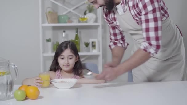 Šťastný blízkovýchodní otec v zástěře dávat pomerančový džus roztomilé dceři jíst cereálie ráno. Mladý muž líbá holčičku do hlavy a usmívá se. Péče, jednota, rodina, životní styl. — Stock video