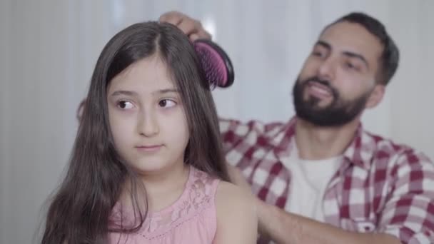 Jonge lachende man uit het Midden-Oosten kamt lang zwart haar van dochter uit. Een oplettende vader die thuis voor een leuk klein meisje zorgt. Liefde, familie, vaderschap, levensstijl. — Stockvideo