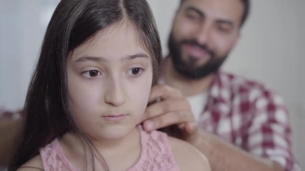 Close-up face of beautiful little girl with brown eyes standing indoors as blurred man combing her long black hair at the background. Middle Eastern father taking care of pretty daughter. — Stock Video