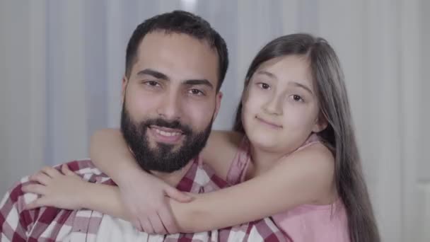 Närbild porträtt av glad Mellanöstern far och dotter ler mot kameran. Brun-ögd man och liten flicka med svart hår tillbringar tid hemma. Lycka, enhet, livsstil. — Stockvideo