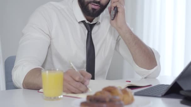Giovane bell'uomo d'affari mediorientale che parla al telefono e fa colazione al mattino. Uomo impegnato in camicia bianca e cravatta nera avendo spuntino. Multitasking, business, lifestyle . — Video Stock