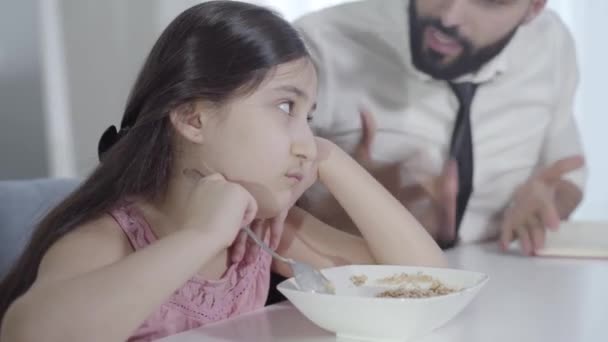 Sfocato giovane uomo del Medio Oriente urlando triste ragazzina mescolando porridge con cucchiaio. Ritratto di padre affrettato che litiga con figlia disobbediente al mattino. Stile di vita, colazione, genitorialità . — Video Stock
