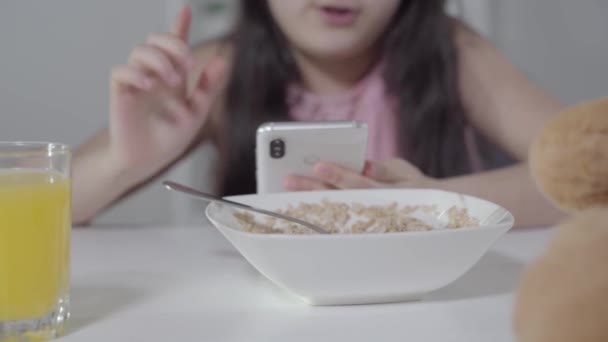 Primo piano di piatto con porridge in piedi sul tavolo come sfocata bambina carina utilizzando smartphone sullo sfondo. Abbastanza bambino mediorientale che usa i social media durante la colazione. Stile di vita . — Video Stock