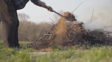Olgun bir Kafkas çiftçisi, kuru yaprakları ve dalları kürekle ateşe atıyor. Çevresel sorunlar, atık yakma karşıtı ekolojik yollar.