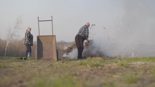 Breed schot van volwassen blanke man die gevallen bladeren in brand gooit als zijn oudere vrouw aan de kant. Plattelandsboeren die droge takken verbranden. Anti-ecologische levensstijl, milieubescherming. — Stockvideo