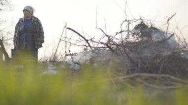 Arka planda yanan ateşin yanında duran kıdemli beyaz kadın. Dişi çiftçi, açık havada kurumuş yaprakları ve dalları yakıyor. Anti-ekolojik yakma, kırsal yaşam tarzı.