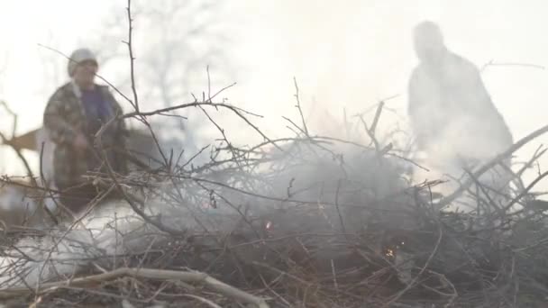 Egy rakás száraz ág égett az előtérben, mint idős pár kaukázusi farmer, akik a háttérben irányítják a tüzet. Öreg férfi és nő elégeti a hulladékot a szabadban. Antiökológiai életmód. — Stock videók
