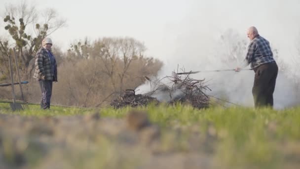 Grande tiro do velho agricultor caucasiano usando ancinho para misturar o fogo da queima de galhos secos. Homens e mulheres mais velhos a incinerar resíduos ao ar livre na Primavera. Estilo de vida, danos ambientais, poluição . — Vídeo de Stock