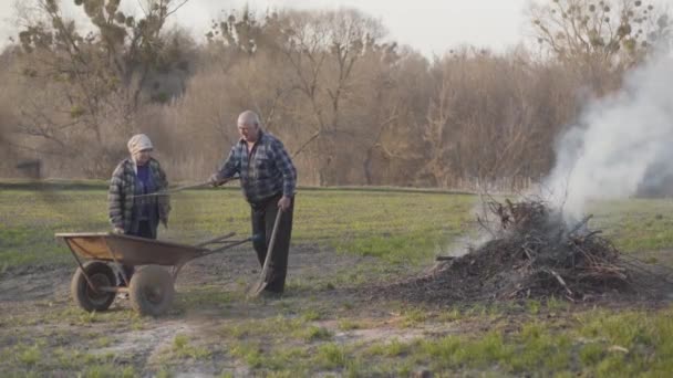 Een paar volwassen blanke boeren verzamelen tuingereedschap en laten open haard met droge takken branden. Verbranding van afval, milieuschade en verontreiniging. — Stockvideo