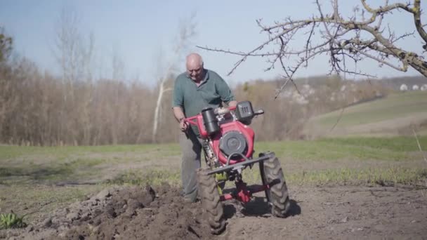 Ritratto di anziano caucasico che cammina lungo il giardino rurale con furrower. Vecchio agricoltore solcare terreno fertile nella soleggiata giornata primaverile. Coltivazione, agricoltura, agricoltura, lifestyle . — Video Stock