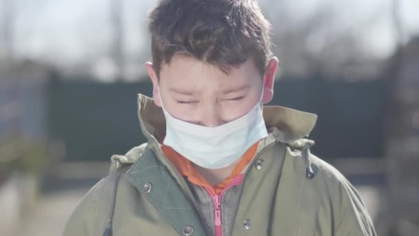 Nahaufnahme eines niedlichen brünetten Kaukasischen Jungen mit schützender Gesichtsmaske, der niest. Kleines braunäugiges Kind, das draußen in der Sonne steht und in die Kamera schaut. Virusinfektion, Covid-19, Träger, Gesundheitsfürsorge. — Stockvideo