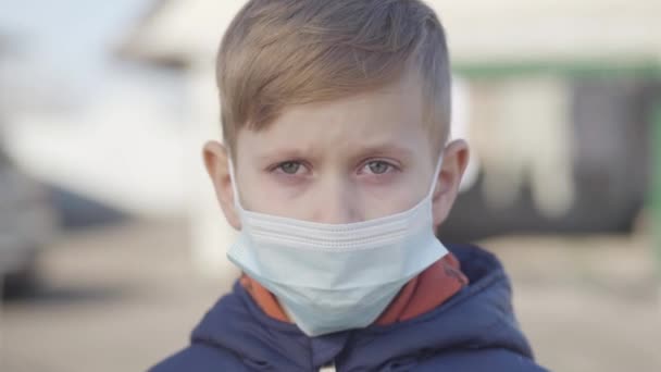 Portrait of cute blond Caucasian boy in protective face mask posing outdoors on sunny spring day. Serious ill child with red eyes looking at camera. Covid-19, global pandemic, coronavirus. — Stock Video