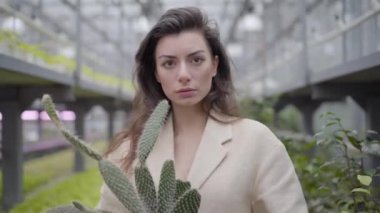 Kaktüs tutan ve kameraya bakan zarif bej ceketli esmer kadının portresi. Cazibeli beyaz kadın cam evde dikiliyor..