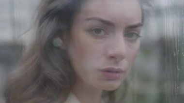 Güzel beyaz esmer kadın yağmurlu bir günde pencereden kameraya bakıyor. Camın arkasında kahverengi gözlü yalnız bir kadının portresi. Kamera soldan sağa hareket ediyor.