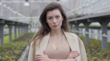 Yetişkin, kahverengi gözlü, serada poz veren beyaz bir kadın. Glasshouse 'da kameraya bakan çekici esmer kız. Kamera soldan sağa hareket ediyor.
