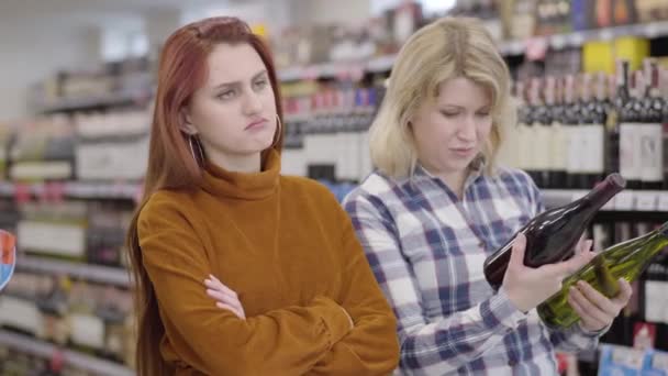 Verveelde roodharige blanke vrouw wachtend op vrouwelijke vriend om te kiezen tussen rode en witte wijn in de winkel. Jonge charmante meisjes die drank kopen in de supermarkt. Lifestyle, vrije tijd, vriendschap. — Stockvideo