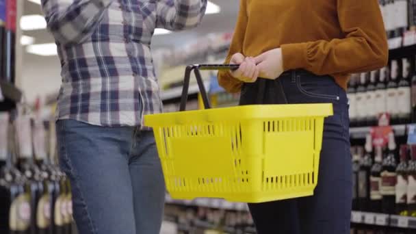 スーパーでアルコールを入れた棚の間に立つ2人の有名な白人女性と、赤ワインと白ワインのボトルをショッピングバスケットに入れる。大人の女の子が店で飲料を買う。ライフスタイル. — ストック動画