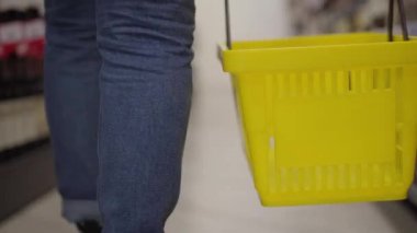 Kot pantolonlu, sarı alışveriş sepetiyle süpermarket raflarında yürüyen, tanınmayan birinin yakın çekimi. Şarap dükkanındaki müşteriyi takip eden kamera. Tüketim, hipermarket, yaşam tarzı.