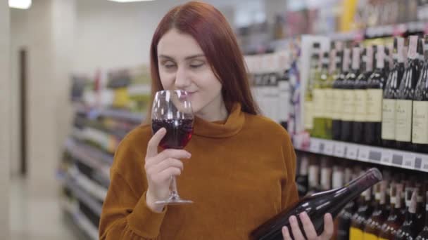 Porträtt av charmig rödhårig kaukasisk kvinna som luktar och förtalar rött vin från glas och gestikulerar ja genom att skaka på huvudet. Ung flicka provsmakning dryck i alkoholbutik. Livsstil, glädje, val. — Stockvideo