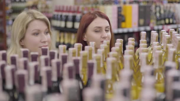 Mujer pelirroja joven con ojos verdes mirando por encima de los estantes con alcohol como alegre amigo rubio unirse a ella. Felices amigas adultas eligiendo bebidas en la tienda de licores. Alegría, ocio, estilo de vida . — Vídeos de Stock