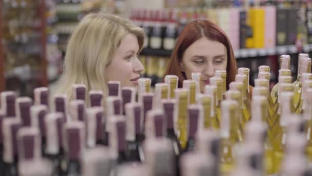 Rostros de dos encantadoras jóvenes caucásicas detrás de botellas de alcohol en el estante. Mujeres amigas positivas charlando como la selección de vino en la tienda. Alegría, ocio, estilo de vida, diversión . — Vídeos de Stock