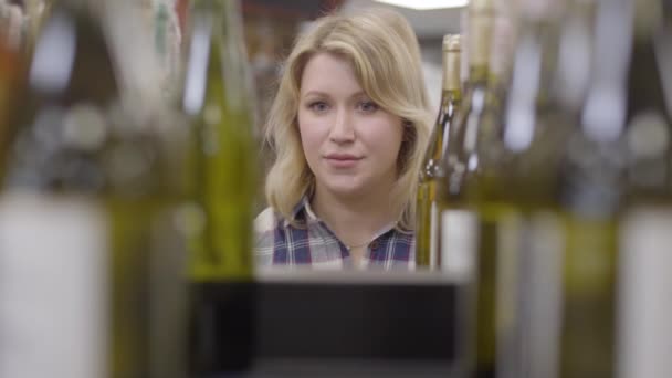 Rostro de mujer rubia caucásica positiva que aparece entre botellas de vino blanco, sonriente dama tomando una bebida y marchándose. Jovencita alegre comprando bebida en el supermercado de alcohol . — Vídeo de stock