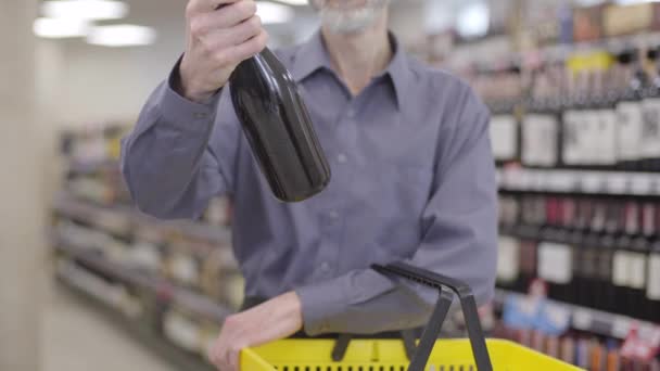 Oigenkännlig skäggig senior man tittar på vin flaska och sätta den i gul kundvagn. Manlig kund som köper dyr dryck i en alkoholbutik. Konsumtion, alkoholism, livsstil. — Stockvideo