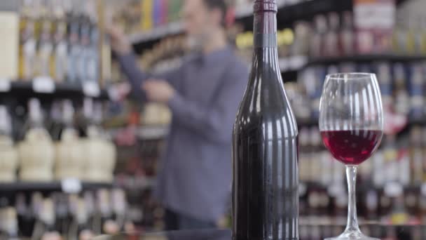 Detailní záběr na láhev vína a sklenici s nápojem stojící v popředí s rozmazaným bělochem, který si v pozadí vybírá nápoj. Supermarket s alkoholem, životní styl, degustace. — Stock video