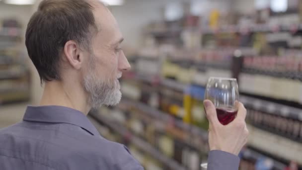 Vista laterale primo piano del sommelier caucasico adulto che scuote e annusa il vino rosso nel bicchiere. Sorridente steward professionista del vino che verifica la qualità delle bevande nel negozio di liquori. Stile di vita, perfezione, alcol . — Video Stock