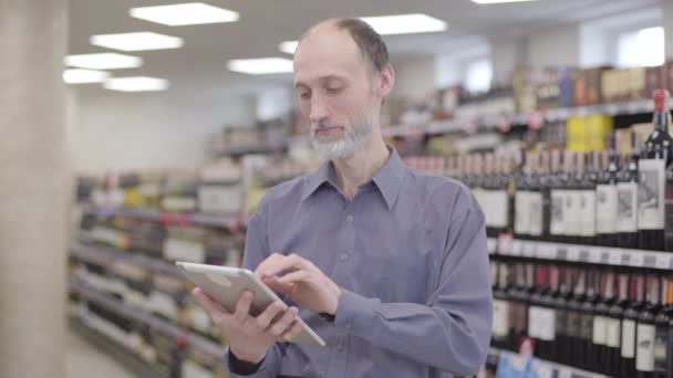 Portret van professionele senior handelaar met behulp van tablet in een alcoholsupermarkt. Zelfverzekerde blanke man met baard die het scherm veegt en lachend tussen rijen dranken in de winkel staat. — Stockvideo
