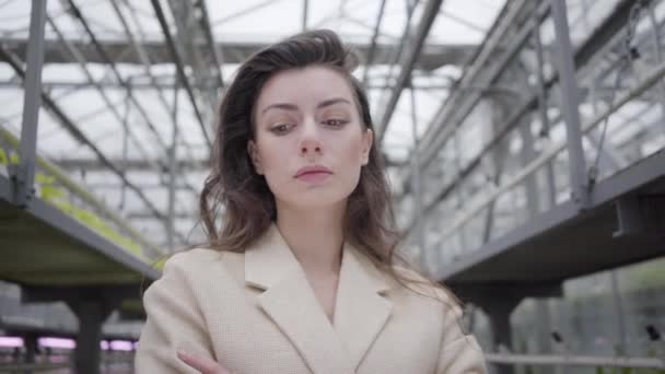 Thoughtful enigmatic woman standing in greenhouse. Camera moving away from Gorgeous young brunette lady looking at camera in glasshouse. — Stock Video