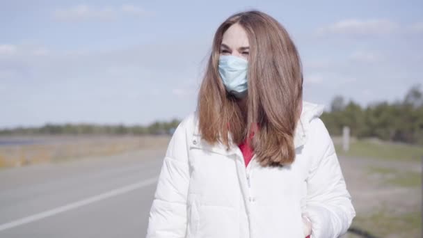 Portret van een jonge blanke vrouw met een beschermend gezichtsmasker op een lege voorstedelijke weg. Mooi brunette meisje brengen zonnige dag buiten tijdens quarantaine. Covid-19, coronavirus, pandemie. — Stockvideo