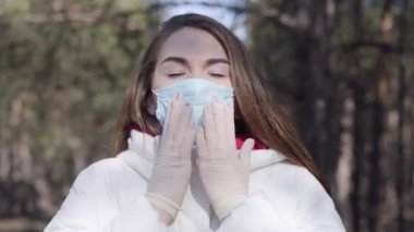 Güneşli bahar parkında öksüren koruyucu maskeli ve eldivenli esmer kadının yakın çekimi. Kafkasyalı hasta kızın portresi koronavirüs hastalığı belirtileri gösteriyor. Covid-19 salgını, yaşam tarzı.