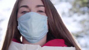 Koruyucu maskeli güzel beyaz kızın yakın plan yüzü. Covid-19 karantinası sırasında güneşli bir bahar gününde dışarıda poz veren güzel esmer kadın. Koronavirüs, küresel salgın, bulaşıcı hastalıklar.