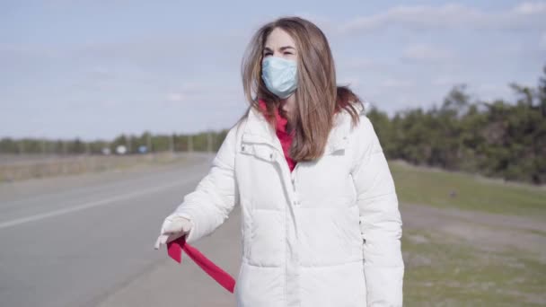 Porträtt av vacker brunett vit kvinna i skyddande ansiktsmask liftar på tom förort väg. Problem med karantän för coronavirus. Covid-19, pandemi, livsstil. — Stockvideo