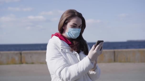 Giovane bruna donna caucasica in maschera facciale e guanti protettivi utilizzando smartphone. Ragazza carina a riposo su imbarco urbano in giornata di sole da solo. Coronavirus quarantena, Covid-19, tempo libero all'aperto . — Video Stock