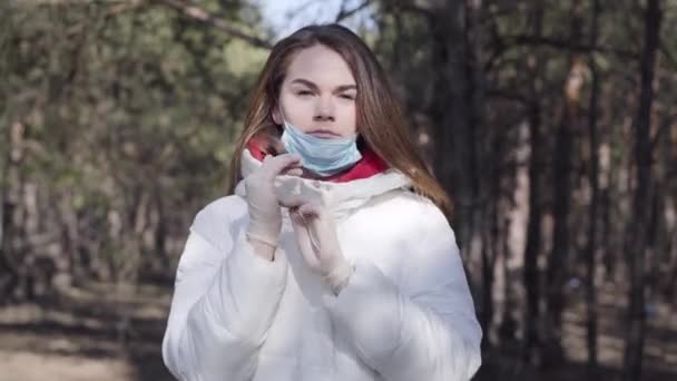 Pewna siebie biała kobieta zakładająca rękawiczki ochronne i maskę. Portret młodej brunetki w ubraniu ochronnym podczas kwarantanny Covid-19. Koronawirus, bezpieczeństwo, styl życia. — Wideo stockowe