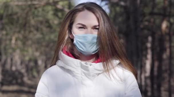 Retrato de cerca de una mujer caucásica morena de ojos grises con mascarilla protectora al aire libre. Niña segura de pasar el soleado día de primavera en el parque o el bosque. Covid-19, pandemia, estilo de vida . — Vídeo de stock