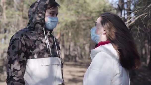 Vista laterale di positivo giovane coppia caucasica in maschere facciali parlando all'aperto. Uomo e donna che trascorrono una soleggiata giornata primaverile al parco durante la quarantena del coronavirus. Covid-19, stile di vita, pandemia, pericolo . — Video Stock