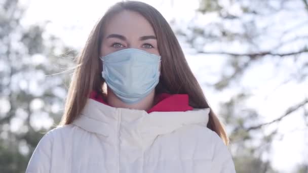 陽射しの中屋外に立つ顔マスクの若い女性。日当たりの良い公園でコロナウイルスの隔離中に保護服を着ているブルネットの白人の少女。Covid-19パンデミック感染症. — ストック動画