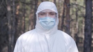 Güneşli bahar parkında duran koruyucu beyaz elbiseli, gözlüklü ve maske takan beyaz bir gencin portresi. Koronavirüs salgını tehlikesi kavramı, karantina, koruma tedbirleri.