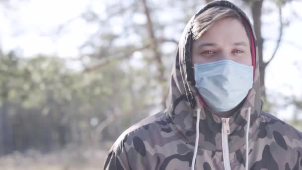 Retrato de niño caucásico con mascarilla protectora de pie en el soleado parque de primavera o bosque y mirando a la cámara. Joven posando al aire libre durante la cuarentena de Covid-19. Coronavirus, virus infeccioso . — Vídeo de stock