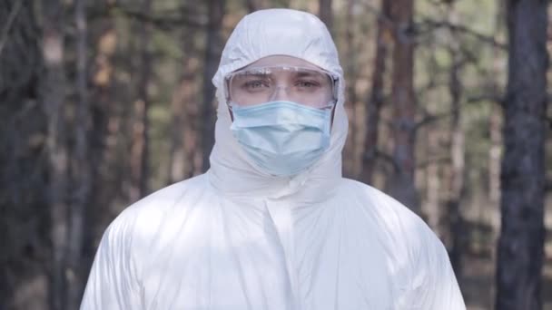 Porträt eines jungen kaukasischen Mannes in weißer Schutzkleidung, Brille und Gesichtsmaske, der im sonnigen Frühlingspark steht. Konzept der Gefahr einer Coronavirus-Pandemie, Quarantäne, Schutzmaßnahmen. — Stockvideo