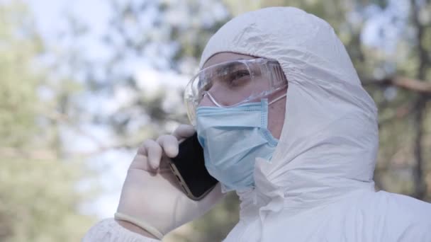 Vue latérale rapprochée du jeune homme caucasien en combinaison de sécurité, lunettes de protection et masque facial parlant au téléphone à l'extérieur. Un gars confiant qui passe le printemps dans un parc ensoleillé. Covid-19 quarantaine . — Video